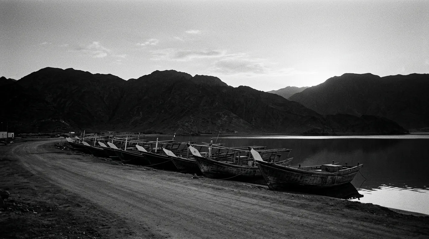Port de pêche de Dibba à l'aube, barques en bois colorées alignées sur une eau plate, mer calme du golfe d'Oman, montagnes Hajar noires en arrière-plan, rendu presque monochrome en teintes graphite