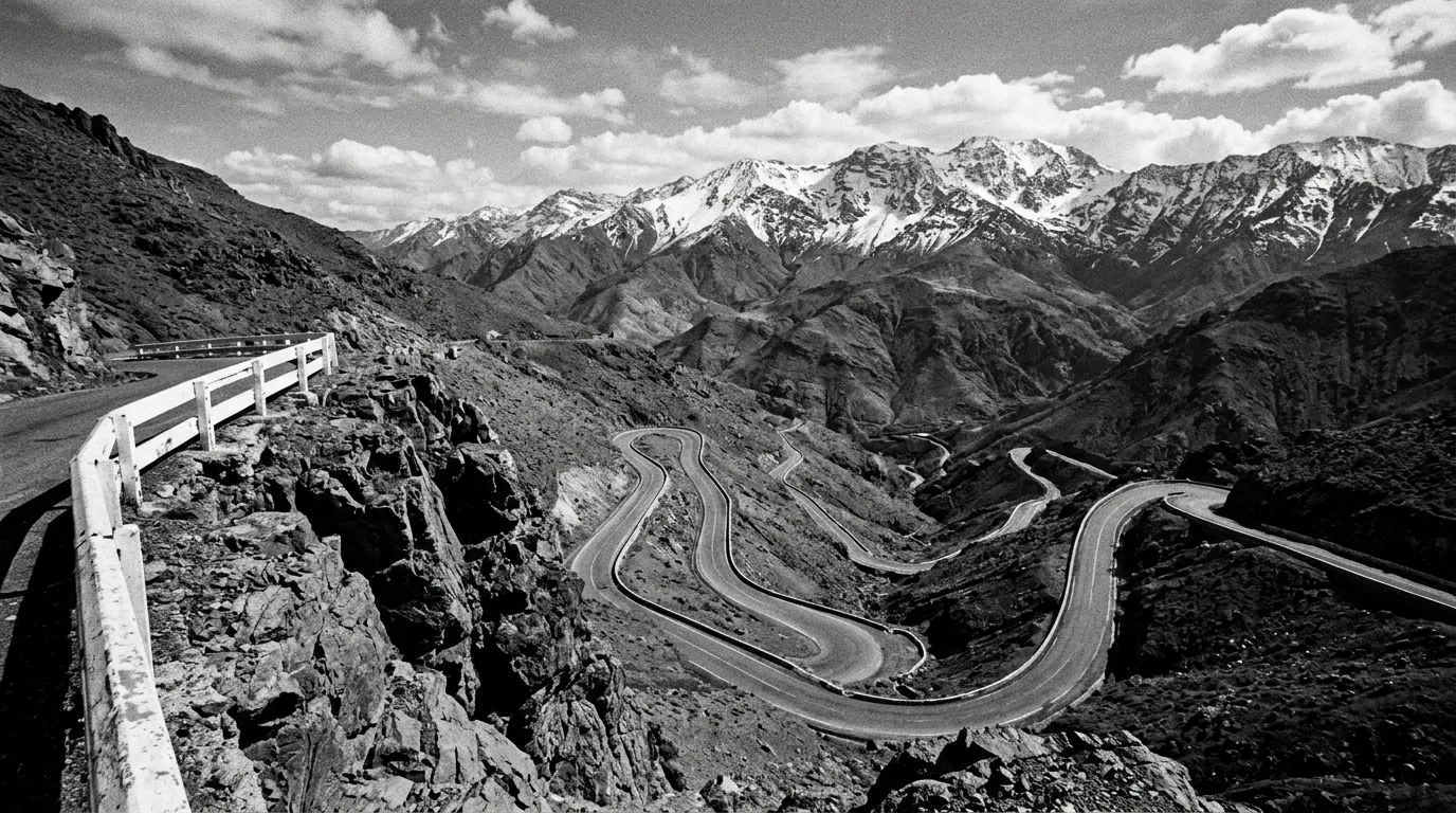 Virages en épingle du col du Tichka vus depuis un éperon rocheux, barrière blanche de sécurité, route N9 qui serpente en lacets serrés, cimes enneigées de l'Atlas au fond