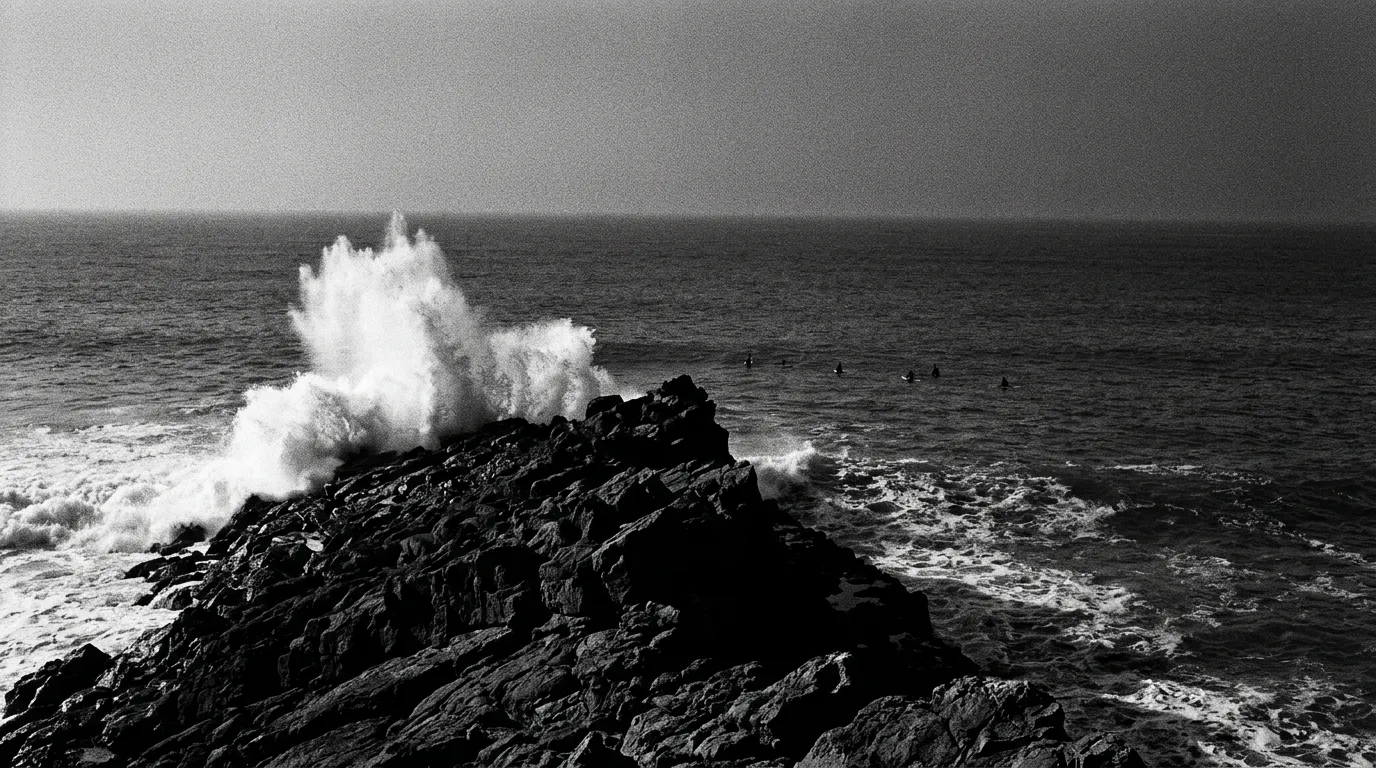 Pointe rocheuse au sud de Taghazout en fin d'après-midi, écume blanche qui explose sur un basalte noir, silhouettes minuscules de surfeurs au lineup qui attendent une série