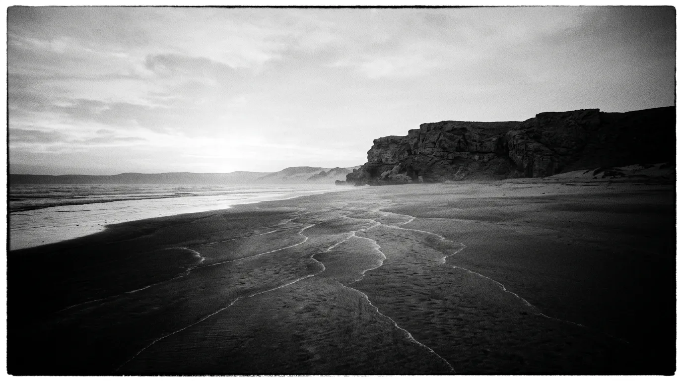 Plage de Ras al-Jinz au lever du jour, lumière basse rasante, sable humide avec traces ondulées remontant de la mer vers les dunes, falaise rocheuse au fond, aucune présence humaine