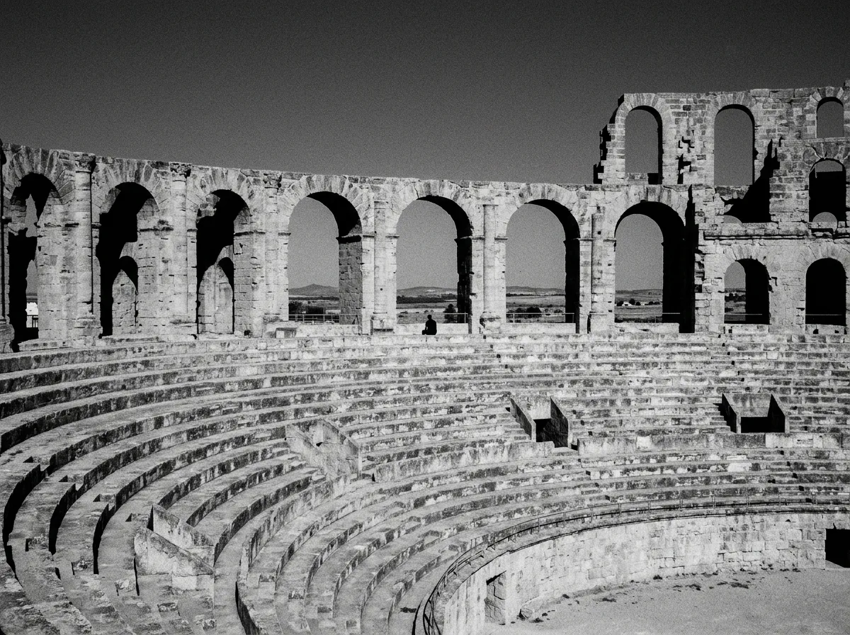 Arcades et gradins de l'amphithéâtre d'El Jem vus depuis l'intérieur de l'arène, pierre dorée au soleil bas, gradins presque déserts, ciel bleu profond